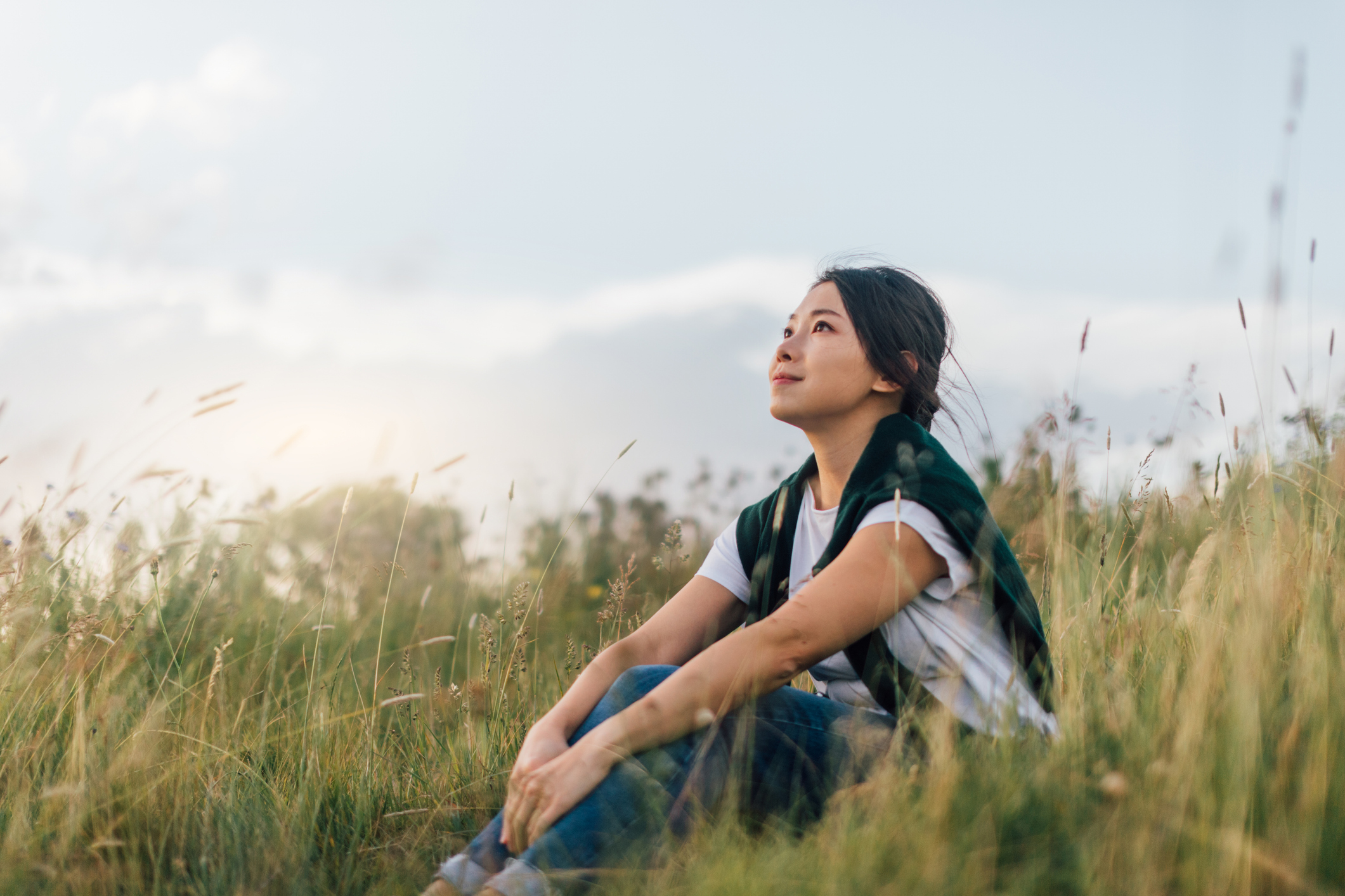 young woman sitting in nature with blue shirt on and sweater wrapper around her shoulders. grass is green and sky is blue