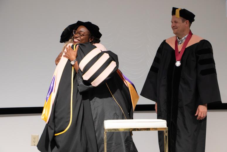 Graduate received their hood and is hugging a faculty member smiling while another faculty member is smiling next to them. All wearing regalia