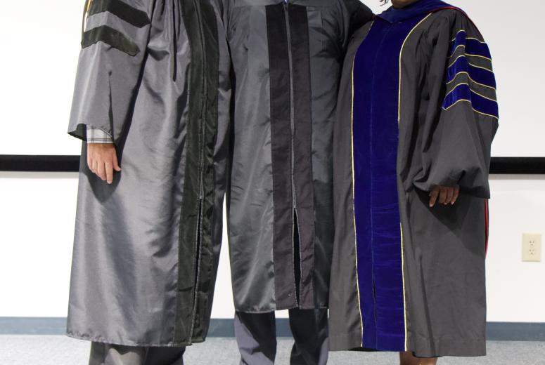 3 individuals standing on a stage in regalia looking at the camera and smiling in celebration of the hooding ceremony