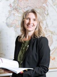 Anna Nelson smiling holding a book in front of a map with a black blazer and sage green shirt