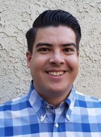 Alan Cupino smiling with plaid blue and white shirt behind a white wall