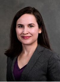 Laura Evans wearing a gray blazer with purple undershirt smiling in front of a gray backdrop.