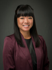 Esther Guerpo smiling with a grayish blackish background and a purple blazer with a black undershirt.