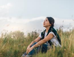 young woman sitting in nature with blue shirt on and sweater wrapper around her shoulders. grass is green and sky is blue